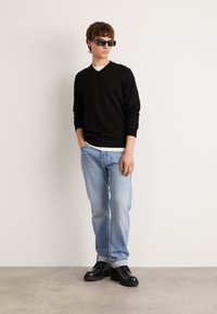 Schwarzer V-Ausschnitt-Pullover, hellblaue verwaschene Jeans und schwarze Schnürstiefel. Die Accessoires umfassen schwarze übergroße Sonnenbrillen. Texturierte Materialien.