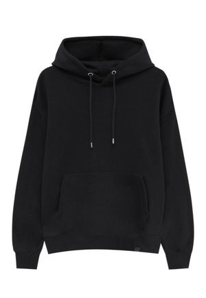 Person trägt einen schwarzen Hoodie mit Fronttasche und weißen Kordeln, kombiniert mit locker sitzenden weißen Hosen vor neutralem Hintergrund.