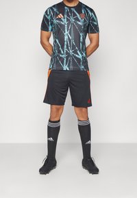 Musta jalkapallopaita turkoosilla geometrisellä kuviolla, oransseilla yksityiskohdilla shortseissa, mustat sukat valkoisilla raidoilla ja mustat nappikset.