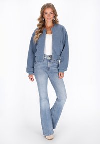 Giacca bomber azzurro chiaro e pelosa con polsini e orlo a costine, indossata sopra una maglietta bianca e jeans chiari a zampa. Design semplice e pulito.