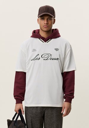 Jeune homme portant une chemise en maille blanche sur un sweat à capuche bordeaux, pantalon gris et casquette marron, tenant un sac sombre dans sa main gauche.