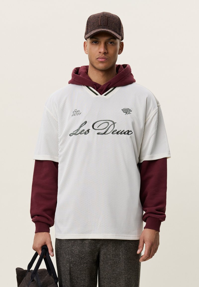 Jeune homme portant une chemise en maille blanche sur un sweat à capuche bordeaux, pantalon gris et casquette marron, tenant un sac sombre dans sa main gauche.