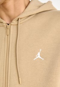 Sudadera beige con cremallera y textura suave. Presenta un logo Jumpman blanco en el pecho y una capucha con cordón de color beige.