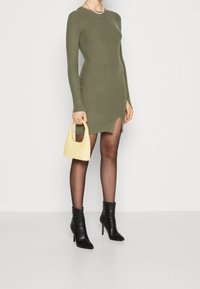 Robe longue à manches longues côtelée olive avec fente sur le côté, associée à un sac à main jaune et des bottes à talons noires. Texture lisse et design ajusté.