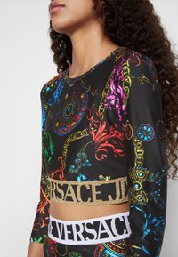 Zwarte cropped top met kleurrijke barokke patronen, voorzien van lange mouwen. Geaccentueerd met een gouden "VERSACE" logo en een witte "VERSACE" band. Glad oppervlak.