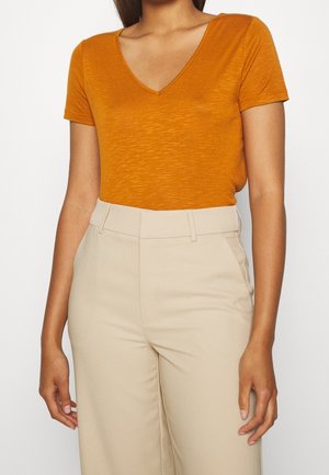 Woman wearing a fitted short-sleeve burnt orange V-neck top and high-waisted beige trousers against a plain background.