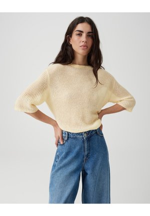 Frau mit langen dunklen Haaren, die einen beigen gestrickten Pullover und lockere blaue Jeans trägt, steht mit den Händen in der Hüfte vor einem schlichten Hintergrund.