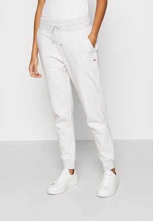 Femme portant un pantalon de jogging gris clair avec taille à cordon et baskets blanches, debout devant un fond blanc uni.