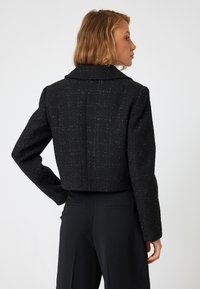 Veste noire courte en tissu texturé, avec un large col et des épaules rembourrées. Le dos est lisse avec une couture centrale.