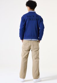 Sudadera azul con "VUELVO EN 2 SEMANAS" impreso en negro en la parte posterior, superpuesta a una camisa a cuadros, combinada con pantalones beige claros y zapatillas blancas.