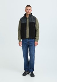 Gilet di pile nero con parte superiore grigia scura e liscia; maglione verde sotto, jeans blu, sneakers nere; vista frontale, modello in piedi.