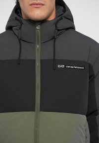 Paprastos striukės su gobtuvu, tamsiai pilkos ir alyvuogių žalios spalvų blokų dizainas. Priekyje zip užtrauktukas ir prekės ženklo detalė krūtinėje.