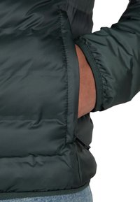 Giubbotto imbottito verde scuro con un esterno testurizzato e imbottito. Presenta polsini elasticizzati e una tasca laterale con zipper. Primo piano della mano in tasca.