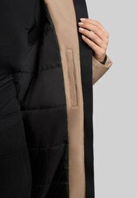 Puffjakke med et beige ydre lag og en sort indvendig. Har en glat tekstur og en sidelomme. Accentdesign langs lukningen.