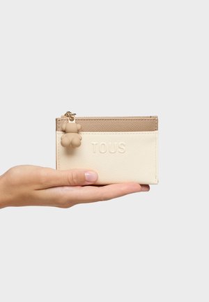 Hand holding beige and cream TOUS cardholder with zipper and small bear keychain charm on white background.