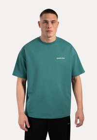 Camiseta oversized en color teal hecha de una tela suave, con un cuello redondo y un logo bordado en blanco en el pecho.