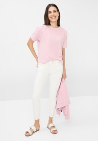 Haut rose clair à manches courtes avec un col arrondi, associé à un jean blanc slim et des sandales claires. Tient une veste rose.
