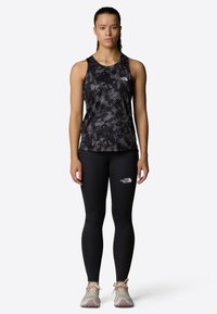 Top a canotta tie-dye nero e grigio, leggings neri aderenti e scarpe da trekking beige. La canotta ha un collo rotondo e dettagli con il logo.