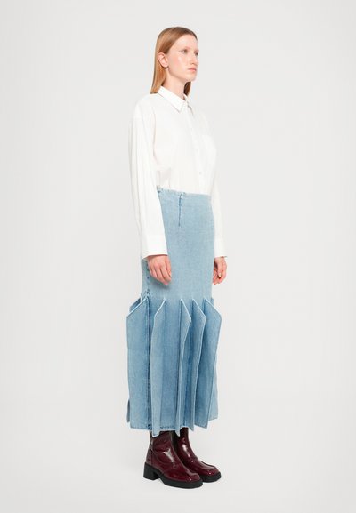 Gonna di denim azzurra chiara con pannelli sovrapposti e frastagliati; indossata con una camicia bianca a bottoni e stivaletti borgogna. Texture liscia.