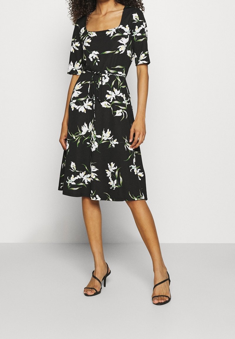 Czarna midi sukienka w kwiaty z kwadratowym dekoltem, krótkimi rękawami i dopasowaną talią. Charakteryzuje się białymi wzorami kwiatowymi i lekko opadającym materiałem.