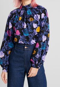 Blouse noire à fleurs avec manches longues, présentant des roses colorées en violet, rose et bleu, un col froncé et un tissu léger.