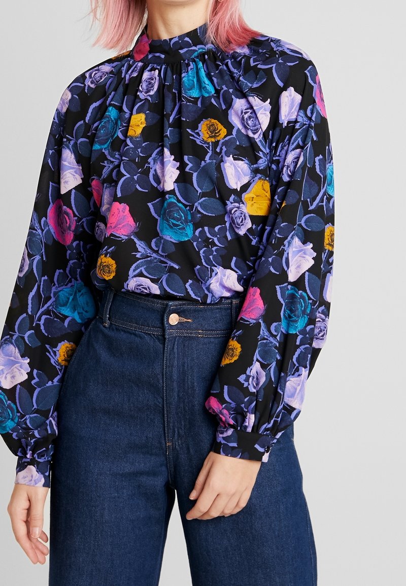 Blouse noire à fleurs avec manches longues, présentant des roses colorées en violet, rose et bleu, un col froncé et un tissu léger.