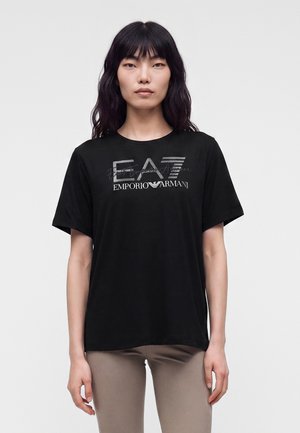 Personne aux longs cheveux noirs porte un t-shirt noir EA7 Emporio Armani et un pantalon beige, debout devant un fond clair uni.