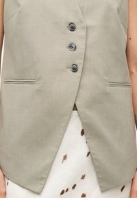 Μπεζ αμάνικο σακάκι με τρία σκούρα κουμπιά και δύο οριζόντιες σχισμές τσέπες, φορεμένο πάνω από μια λευκή φούστα με καφέ πουά.