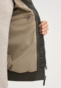 Interno di una giacca imbottita verde con un rivestimento marrone chiaro, caratterizzata da una tasca con zip e orli a costine. Chiusura frontale con zip visibile.