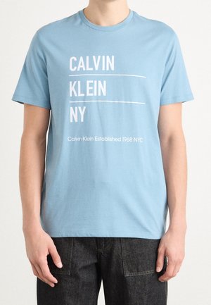Mężczyzna w jasnoniebieskiej koszulce Calvin Klein z białym napisem „CALVIN KLEIN NY” oraz „Calvin Klein Established 1968 NYC”, w połączeniu z ciemnymi dżinsami.