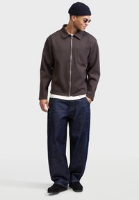 Chaqueta marrón con cremallera, de superficie texturizada y con un bolsillo frontal, combinada con pantalones vaqueros oscuros y sueltos de color azul y zapatos negros. Modelo usando gafas de sol y un gorro de punto.