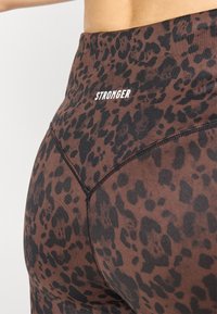Augstās jostasvietas legingi brūnā un melnā leoparda rakstā, ar baltu "STRONGER" logo un redzamām šuvju detaļām.