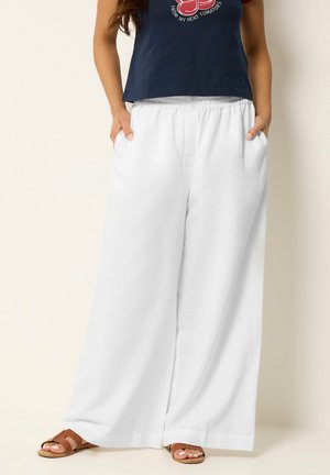 Kobieta ubrana w luźne białe spodnie o szerokich nogawkach z elastycznym pasem i granatowy krótki t-shirt, stojąca z rękami w kieszeniach i brązowych sandałach.