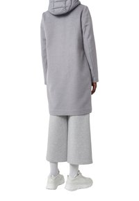 Personne portant un long manteau gris avec capuche, un large pantalon gris court, des chaussettes blanches et de grosses baskets blanches, de dos sur un fond blanc.
