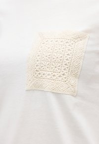 Λευκό ύφασμα με τετράγωνο, υφασμάτινο, κεντημένο μπαλώμα που διαθέτει γεωμετρικά και ανθικά μοτίβα σε ταιριαστή λευκή κλωστή.