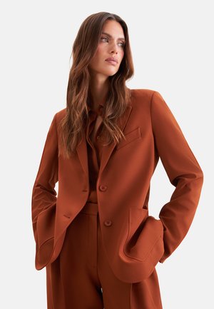 Donna con capelli castani lunghi che indossa un completo su misura color arancione bruciato con le mani in tasca, che guarda di lato su uno sfondo chiaro e uniforme.