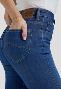 Main reposant dans la poche arrière d'un jean bleu de la marque Lee porté avec un haut blanc, montré de la taille au milieu de la cuisse.