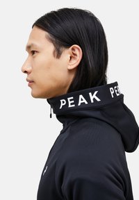 Černá mikina s kapucí a vysokým límcem, na límci je bílý logotyp "PEAK". Hladká textura a atletický střih, ideální pro sportovní nošení.
