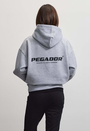 Persona vista da dietro che indossa una felpa grigia con la parola "PEGADOR" e un piccolo testo sotto sulla schiena, abbinata a pantaloni neri.