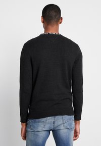 Jersey negro de punto con mangas largas, diseño de escote en pico y dobladillo acanalado. Presenta un ribete de contraste de marca en el escote. Combinado con unos vaqueros de color azul claro.