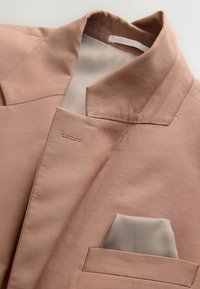 Hellbraune Blazerjacke mit Reverskragen und weißem Einstecktuch in der Brusttasche auf hellem Hintergrund.