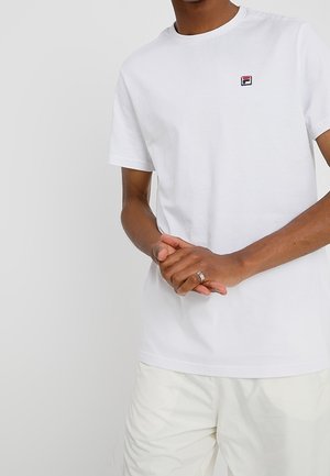 Man draagt een effen wit T-shirt met korte mouwen met een klein rood en zwart logo op de linkerborst, witte broek en een zilveren ring aan de linkerhand.