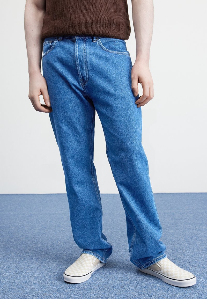 Blue denim jeans with a straight fit, five-pocket design, and subtle fading. Paired with light checkerboard slip-on shoes.