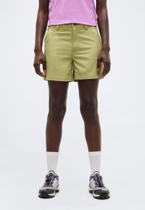 Osoba ubrana w oliwkowe krótkie spodenki, białe skarpetki za kostkę, czarno-białe trampki i różową bluzkę, stojąca na jednolitym tle.