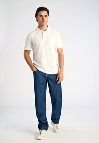 Vit pikétröja med krage, kombinerad med blå jeans i denim. Modell bär vita sneakers och står med handen i fickan mot en ljus bakgrund.