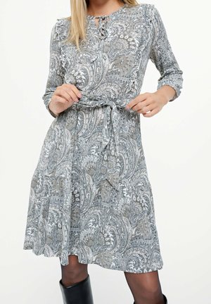 Femme ajustant sa ceinture sur une robe à motif paisley arrivant aux genoux, manches longues et col à nouer, portant des bottes noires et des collants transparents.