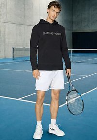 Mladić u crnoj Björn Borg dukserici s kapuljačom i bijelim šorcama drži teniski reket na unutarnjem plavom teniskom terenu.