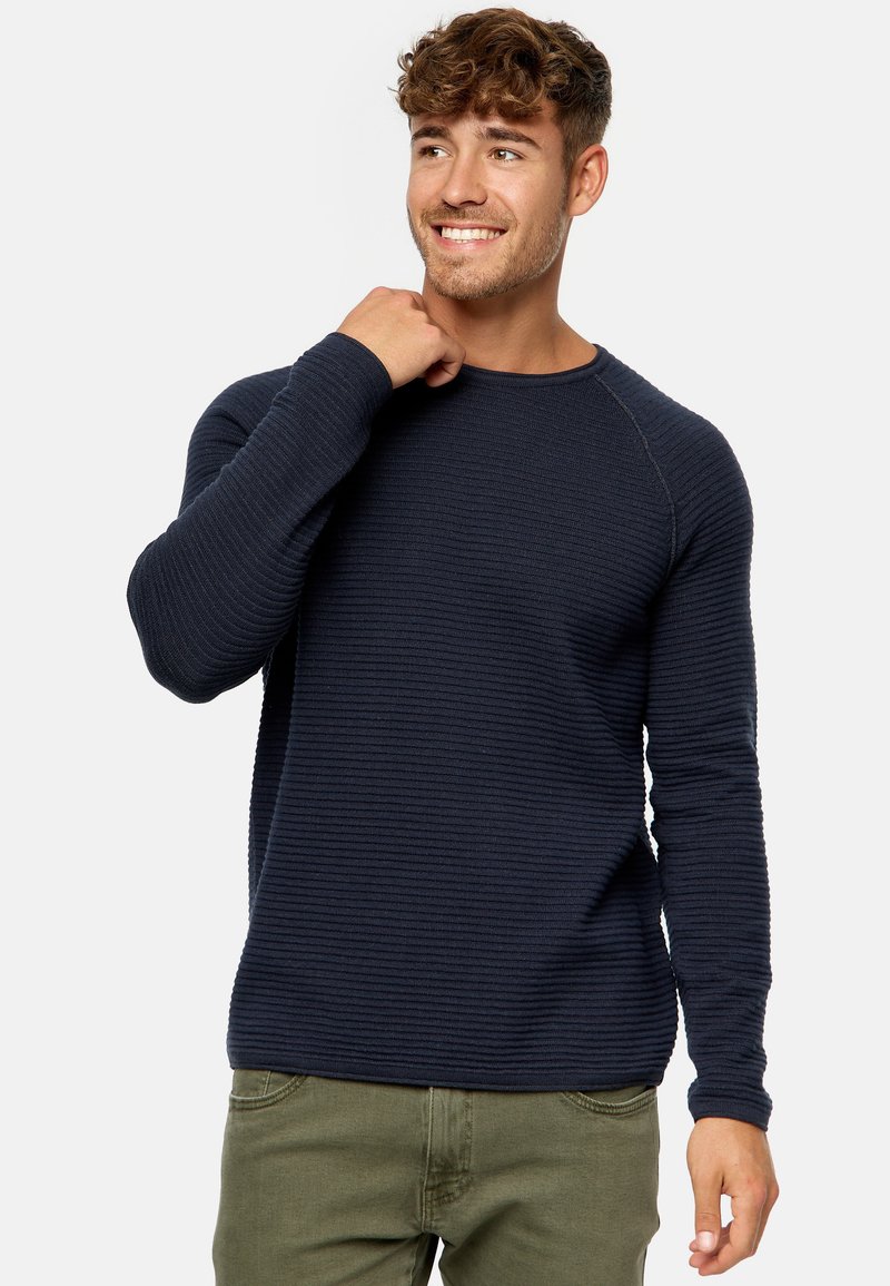 Marineblaue gerippte Pullover mit langen Ärmeln und Rundhalsausschnitt. Ausgestattet mit strukturiertem Stoff und Raglanärmeln, kombiniert mit olivgrünen Hosen.