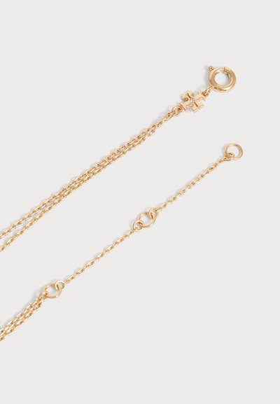 Collier fin en chaîne d’or avec un fermoir circulaire et un petit pendentif en forme de pièce de puzzle proche du fermoir sur un fond clair.