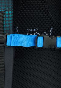 Detail černého síťovaného batohu se modrým nastavitelným popruhem a plastovou sponou, s tečkovaným vzorem, který zdůrazňuje texturu materiálu.